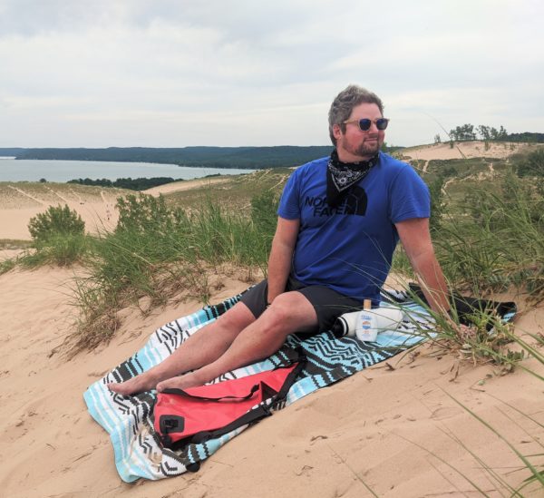Man sitting on beach dunes with recycled Baja Aqua festival blanket, cork sunglasses and reef-safe sunscreen for eco-friendly travel gear