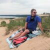 Man sitting on beach dunes with recycled Baja Aqua festival blanket, cork sunglasses and reef-safe sunscreen for eco-friendly travel gear