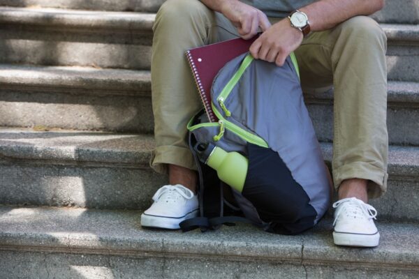 Zoomed in shot of an individual sitting on cement stairs putting away a notebook into their eco friendly Chico Bag brand recycled plastics collapsible travel pack.
