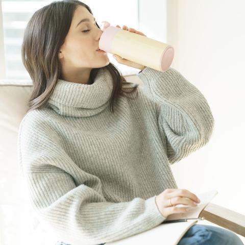 Eco friendly reusable Welly brand Original 12 oz rose colored mug being sipped by female with long brown hair in a comfy turtleneck sweater.