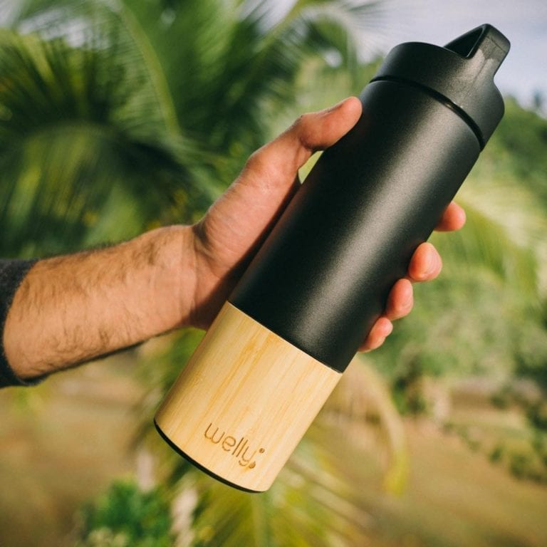 Pictured forearm of male holding a Welly brand 18 ounce black bamboo and stainless insulated traveler bottle loop cap in his hand with palm tree leaves in the background.