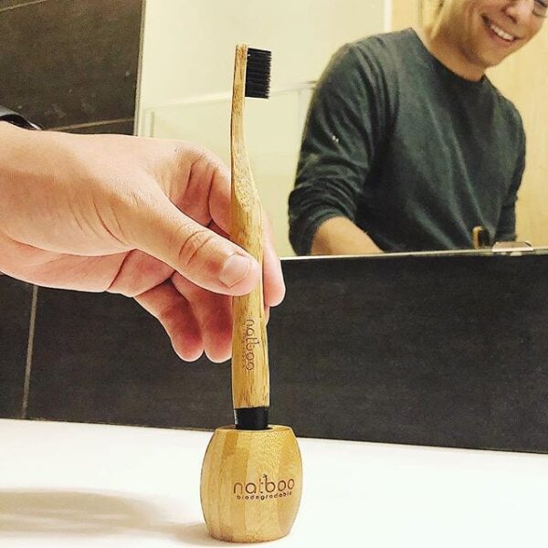 Pictured is a male placing his eco friendly Natboo brand biodegradable round bamboo toothbrush with black tip into small barrel shaped bamboo stand.
