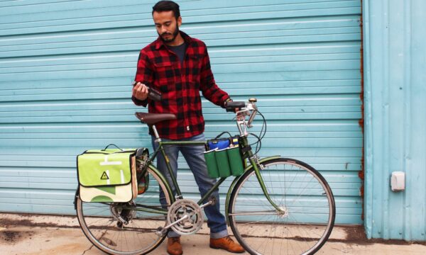 Male standing beside cruiser bike with eco friendly Green Guru Gear brand Sixer 6-pack Top Tube Holder, loaded up with bottle brews, mounted on bike frame.  Product is made from upcycled bicycle inner tubes, repurposed nylon fabric, and 18oz vinyl waterproof tarp.