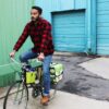 Male in long sleeve red and black flannel riding cruiser bike with eco friendly Green Guru Gear brand Sixer 6-pack Top Tube Holder, loaded up with bottle brews, mounted on bike frame.  Product is made from upcycled bicycle inner tubes, repurposed nylon fabric, and 18oz vinyl waterproof tarp.