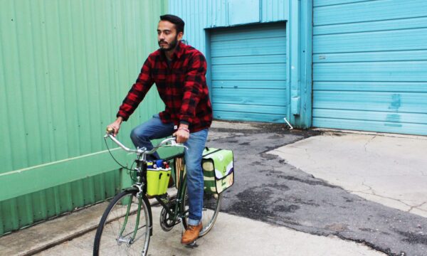 Male in long sleeve red and black flannel riding cruiser bike with eco friendly Green Guru Gear brand Sixer 6-pack Top Tube Holder, loaded up with bottle brews, mounted on bike frame.  Product is made from upcycled bicycle inner tubes, repurposed nylon fabric, and 18oz vinyl waterproof tarp.
