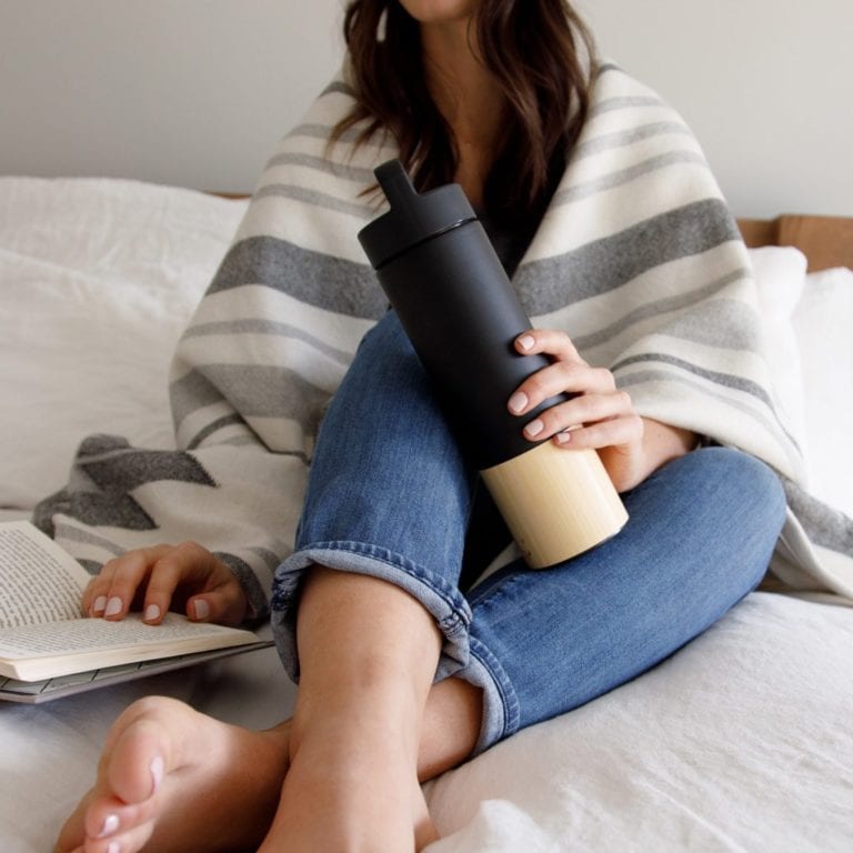 Female sitting in bed reading a book holding a Welly brand 18 ounce black bamboo and stainless insulated traveler bottle loop cap in her hand.