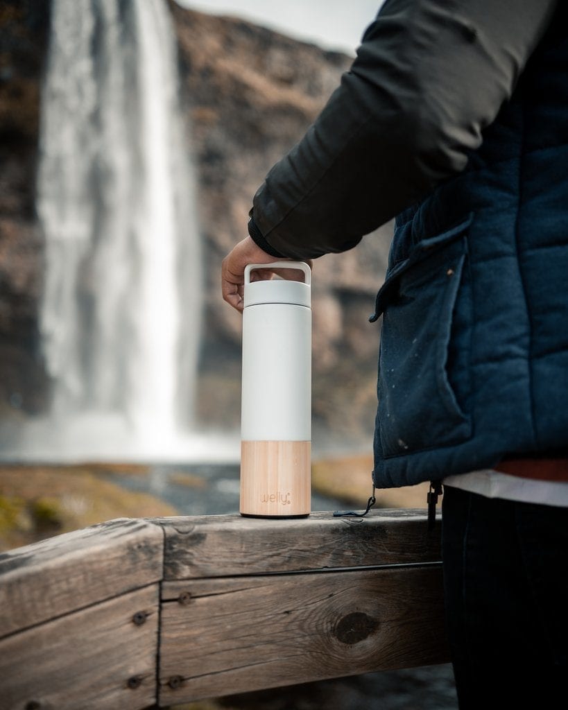 Holding onto loop cap of 18 ounce white bamboo and stainless insulated traveler bottle standing on wood railing with waterfall in the distance.