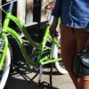 Pictured is a woman standing next to her green cruiser bike holding a sustainably made six liter cruiser cooler handlebar bag made from inner tubers and upcycled material.  Product made by Green Guru Gear brand.