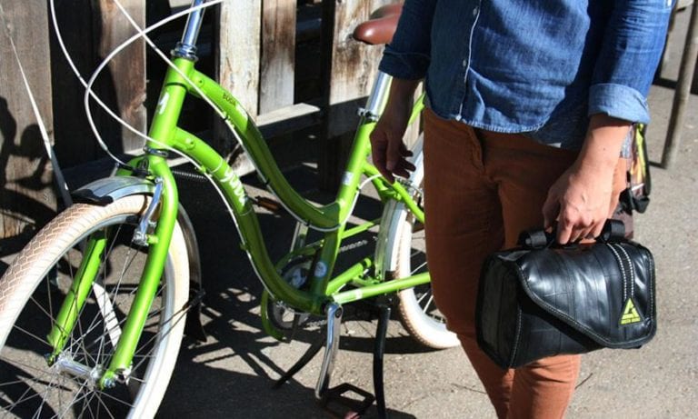 Pictured is a woman standing next to her green cruiser bike holding a sustainably made six liter cruiser cooler handlebar bag made from inner tubers and upcycled material.  Product made by Green Guru Gear brand.