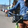 Pictured is a woman standing with her cruiser bike that has a sustainably made six liter cruiser cooler handlebar bag made from inner tubers and upcycled material hanging from chrome handle bars.  Product made by Green Guru Gear brand.