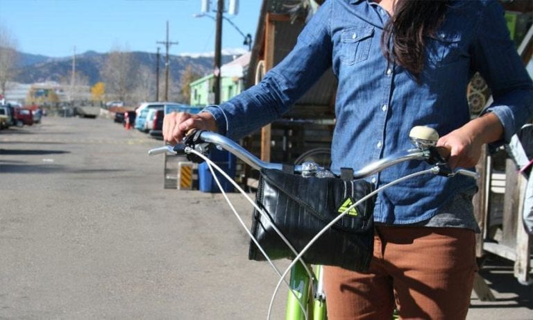 Pictured is a woman standing with her cruiser bike that has a sustainably made six liter cruiser cooler handlebar bag made from inner tubers and upcycled material hanging from chrome handle bars.  Product made by Green Guru Gear brand.