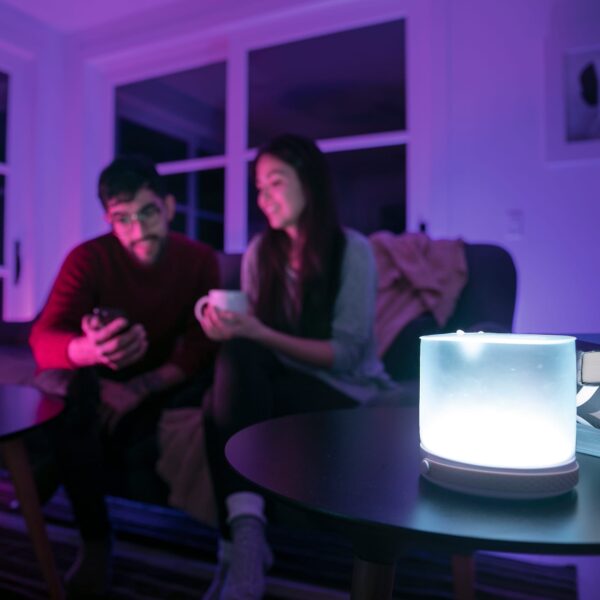 Sustainable Mpowerd brand Luci connect inflatable solar powered light resting on end table inside with color control option used on smartphone app by two people sitting on couch.