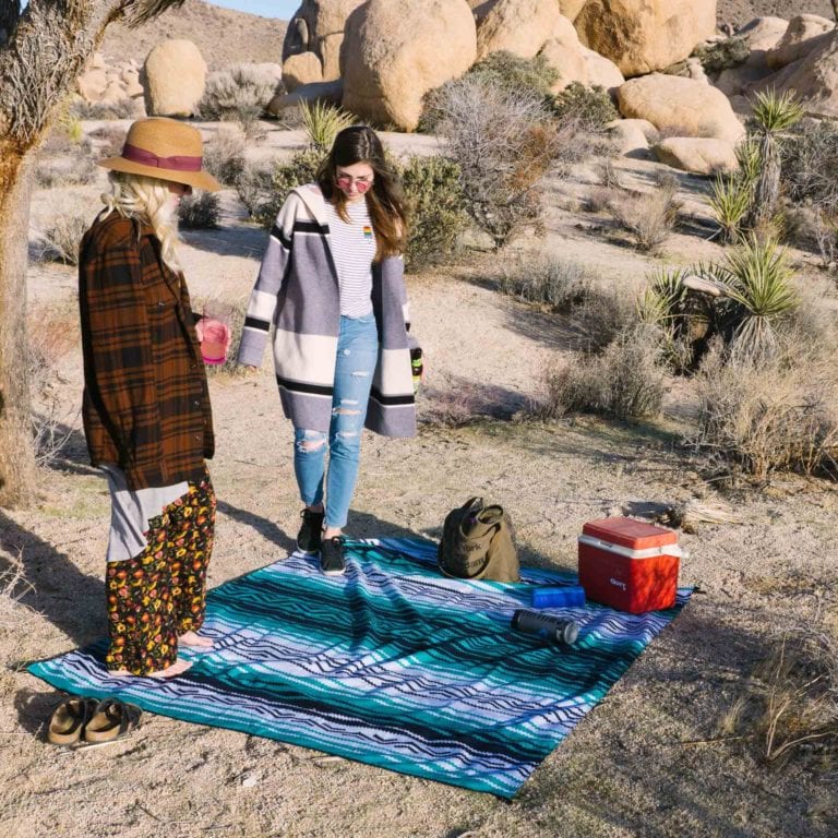 Sustainable Nomadix brand recycled soft and waterproof large Bajaaqua festival blanket spread out in the desert with two females standing on the edges with a red cooler and hiking essentials spread out on the blanket.