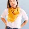 Close up photo of eco friendly gold grid organic cotton square scarf with white tassels, made by Anchal brand, worn loosely around neck of female model.