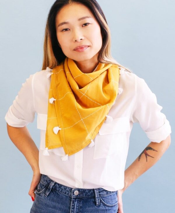 Close up photo of eco friendly gold grid organic cotton square scarf with white tassels, made by Anchal brand, worn loosely around neck of female model.