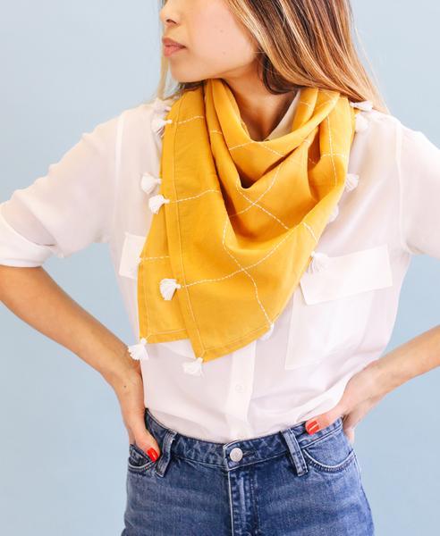 Close up photo of eco friendly gold grid organic cotton square scarf with white tassels, made by Anchal brand, worn loosely around neck of female model.