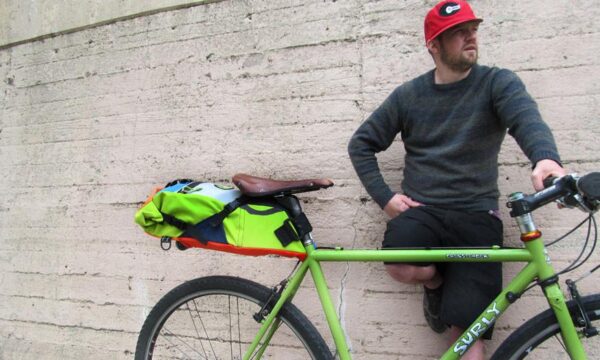 Male posing next to Multicolor upcycled fabric Green Guru Gear brand Haluer Seat Bag Bike Pack displayed in mounted position on light green Surly gravel bike with brown leather seat.
