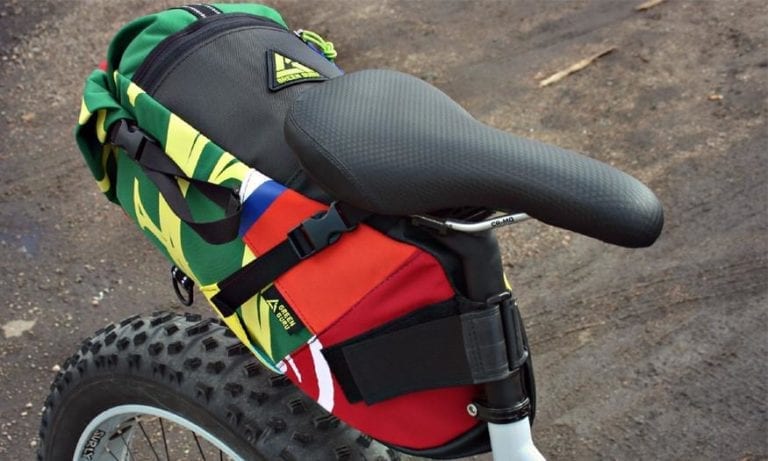 Multicolor upcycled fabric Green Guru Gear brand Haluer Seat Bag Bike Pack displayed on dirt trail mounted on white fat-tire bike with black seat.