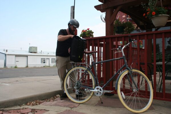 Male removing High Roller 36 liter backpack convertible pannier from rear wheel rack and transition into a backpack.  Pack is made with inner tubes and other upcycled materials by earth friendly Green Guru Gear brand.