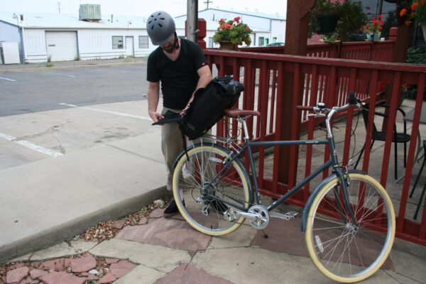 Male removing High Roller 36 liter backpack convertible pannier from rear wheel rack and transition into a backpack.  Pack is made with inner tubes and other upcycled materials by earth friendly Green Guru Gear brand.