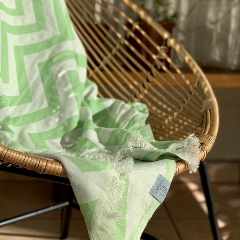 Hilana brand green and white chevron Mersin ultra-soft towel resting on wicker lounging chair, made of sustainable cotton.