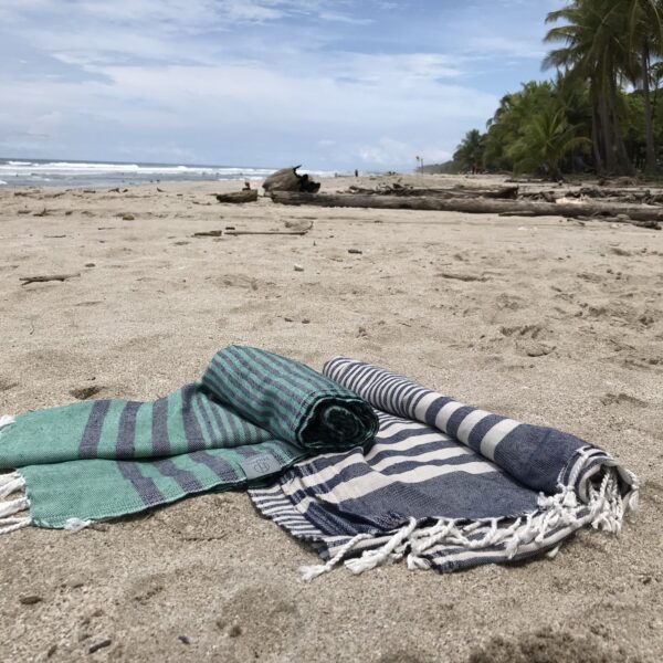 Hilana brand ultra soft navy blue and green and blue striped eco friendly Fethiye towels partially unrolled on the beach.
