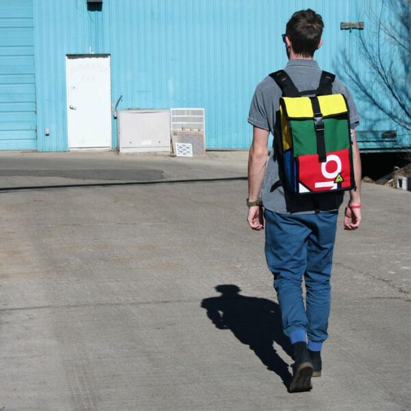 Pictured is a male walking towards blue metal building with back facing the camera wearing an eco friendly Green Guru Gear brand Joyride 24 liter Storm-Proof multi-color Roll Top backpack.