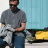 Pictured is a male holding a grey hoodie sitting next to an opened eco friendly Green Guru Gear brand Joyride 24 liter storm-proof multi-color roll top backpack.