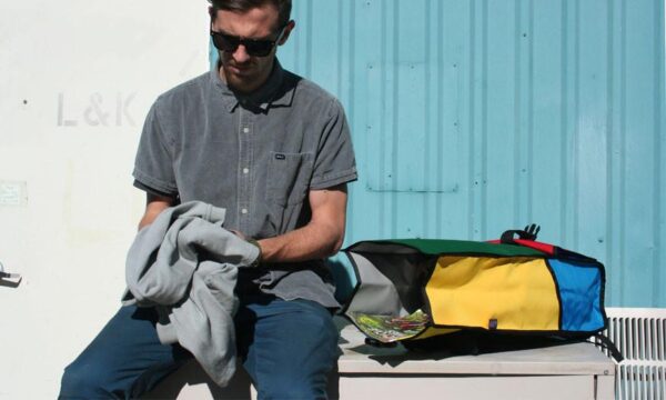 Pictured is a male holding a grey hoodie sitting next to an opened eco friendly Green Guru Gear brand Joyride 24 liter storm-proof multi-color roll top backpack.