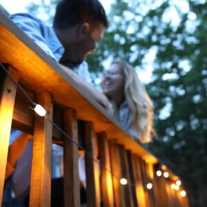 Male and female lean on outdoor deck lit with solar powered stringed white lights, a sustainable product of Mpowerd brand.