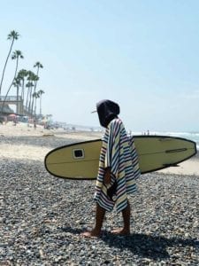 Eco friendly recycled rasta stripe hooded poncho towel sustainably made by Nomadix brand, worn male surfer leaving the beach.