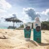 Sustainable All Good Products reef friendly 30 SPF sunscreen spray and lotion nestled into the sand with a tiki umbrella and ocean view in the distance.