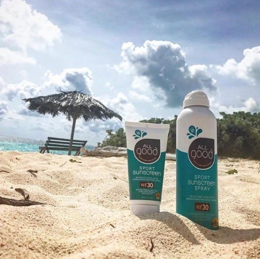 Sustainable All Good Products reef friendly 30 SPF sunscreen spray and lotion nestled into the sand with a tiki umbrella and ocean view in the distance.