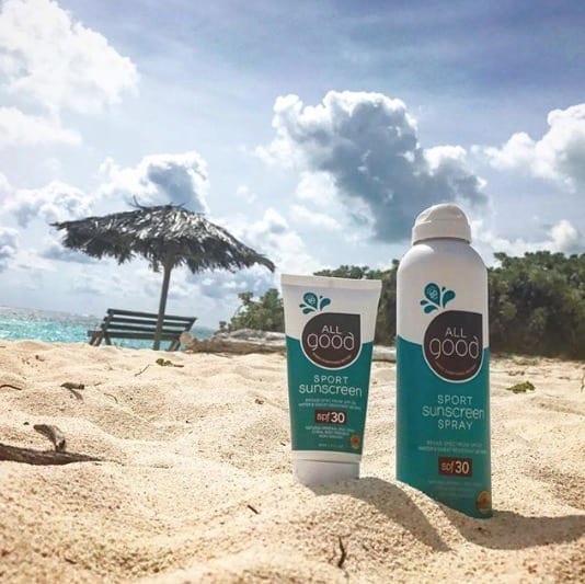Sustainable All Good Products reef friendly 30 SPF sunscreen spray and lotion nestled into the sand with a tiki umbrella and ocean view in the distance.