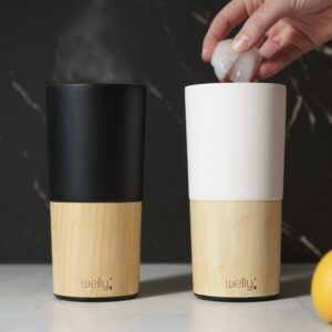 Sustainable Welly brand bamboo and stainless steel insulated 16 ounce black and white tumblers standing side by side.  Hot beverage pictured in black tumbler with ice cubes being added to cold beverage in white tumbler.