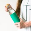Eco friendly Soma brand glass water bottle with emerald green silicone sleeve held in hand of female with the bamboo cap twisted off ready to drink.