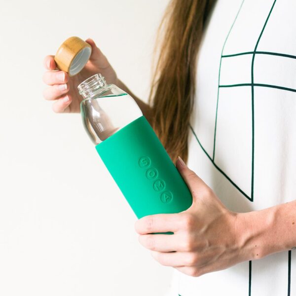 Eco friendly Soma brand glass water bottle with emerald green silicone sleeve held in hand of female with the bamboo cap twisted off ready to drink.