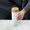 Soma brand eco friendly pearl white insulated ceramic travel mug with bamboo cap in hand displayed on white quartz counter top.