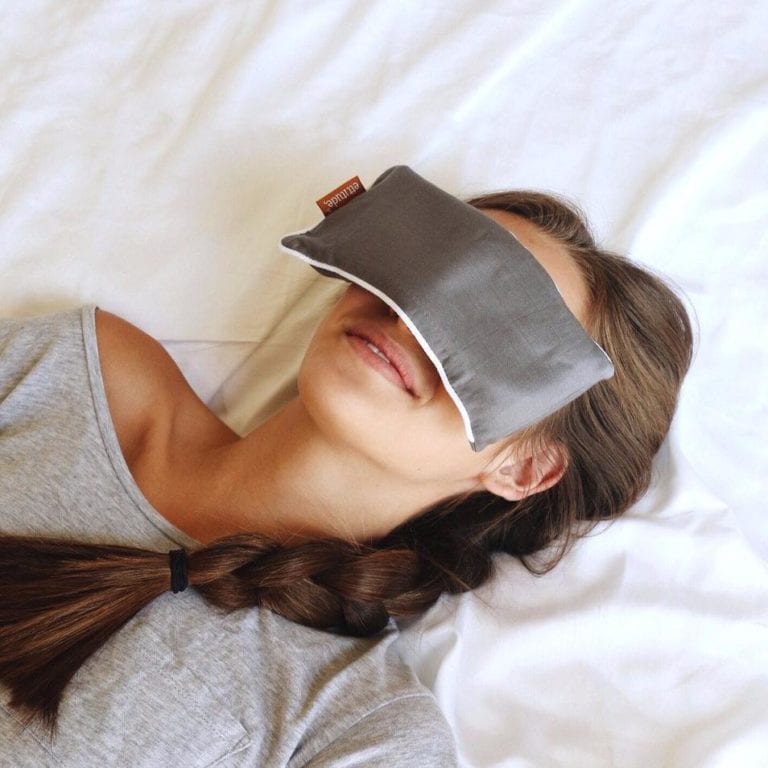 Female with braided long brown hair relaxing in bed with white sheets and a grey organic bamboo eye pillow resting over her eyes. Eye pillow is apart of three piece sleep travel kit made by Ettitude brand.