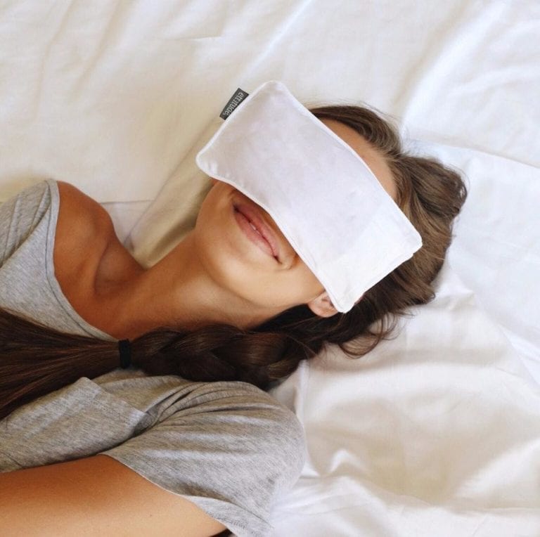 Female with braided long brown hair relaxing in bed with white sheets and a white organic bamboo eye pillow resting over her eyes. Eye pillow is apart of three piece sleep travel kit made by Ettitude brand.