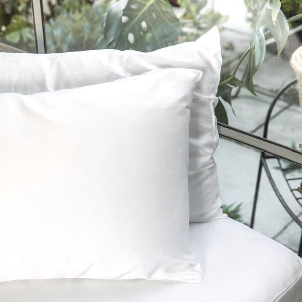 Two standard sized pillows with white organic bamboo pillow cases resting on white ottoman up against window.  One grey organic pillow case is apart of three piece sleep travel kit made by Ettitude brand.