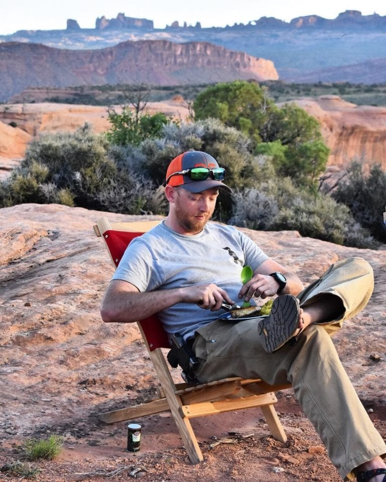 Male sitting outside on rock face eating off reusable dinner plate in a sustainable Blue ridge Chairs brand American ash Blue Ridge Red Camping Chair.