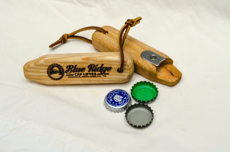 Two sustainable Blue Ridge Chairs brand American Ash upcycled cap lifter bottle opener with three different bottle caps laying in front of cap lifters.