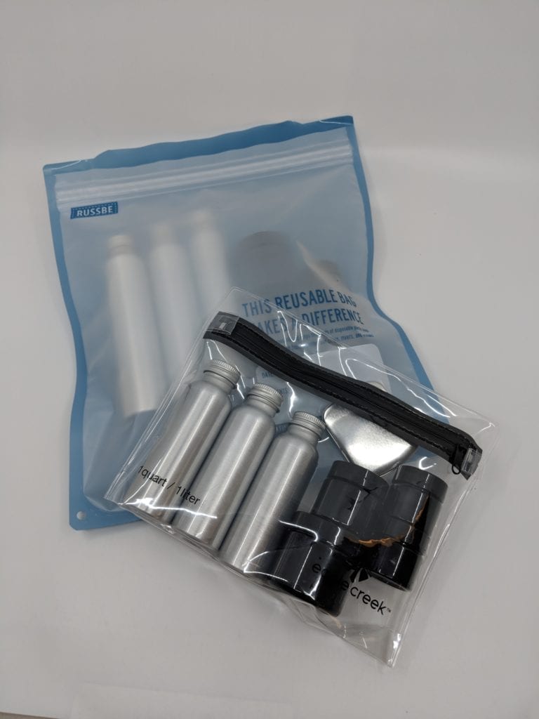 Pictured is part of The Ultimate Reusable Travel Container Set displaying a variety pack of different sized TSA friendly reusable tin containers and plastic containers in clear leak proof bag.