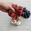 Holding an earth friendly Anchal brand rust scrunchie in hand, made from fair trade organic cotton with a bone and blue colored scrunchies pictured below. Sold together in 3 pack.