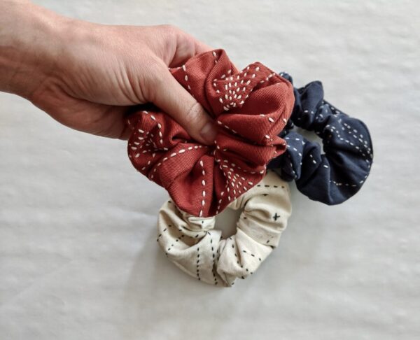 Holding an earth friendly Anchal brand rust scrunchie in hand, made from fair trade organic cotton with a bone and blue colored scrunchies pictured below. Sold together in 3 pack.
