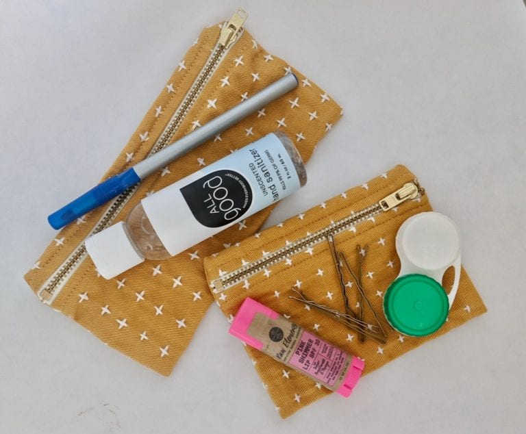 Pen, hand sanitizer, lip balm, contact lenses case and bobby pins spread out across two earth friendly fair trade organic cotton Anchal brand mustard with white plus signs pencil cases.