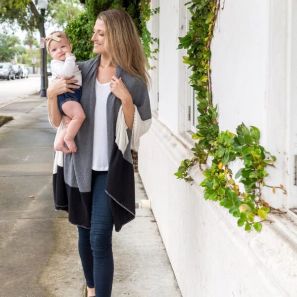 A woman walking her baby down the street while wearing her organic cotton gray colorblock travel shawl. The travel shawl is machine washable for the parent on-the-go.