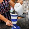 Woman is pouring tea into a grande size Pokito blue reusable cup while in her kitchen.