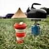 An eco-friendly red Pokito cup is being used while on a camping trip and is sitting in the grass next to a camping stove and tea kettle.